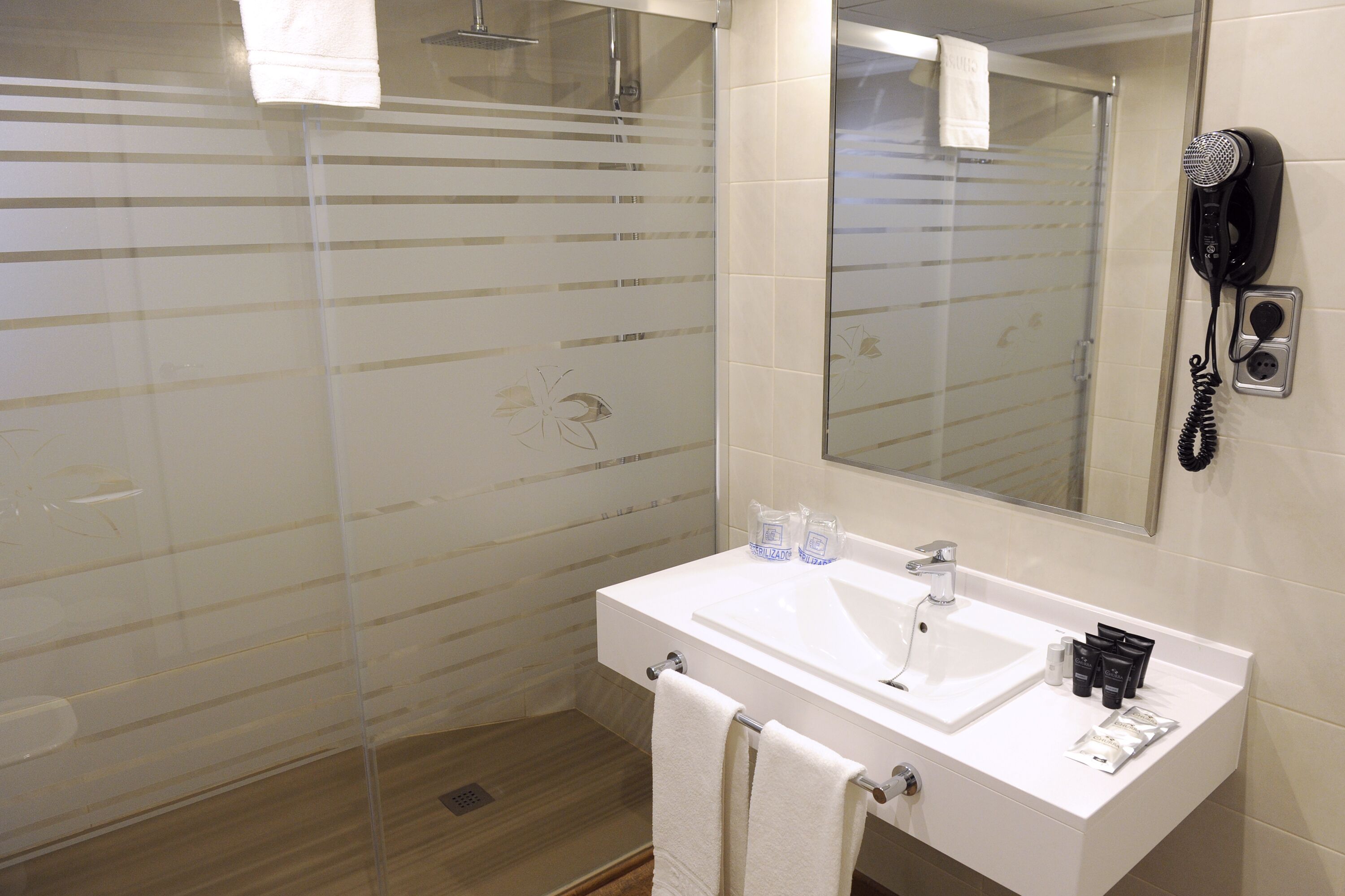 Een moderne badkamer met een glazen douchecabine, een witte wastafel met spiegel en een aan de muur bevestigde haardroger.