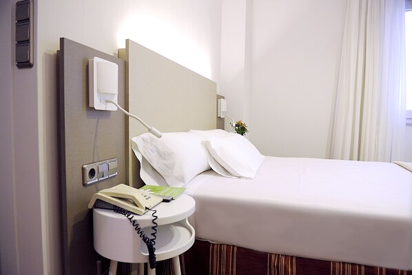 A hotel room with a bed, a bedside table with a telephone and a book, a wall socket, and a window with curtains. - Hotel El Churra (Murcia)