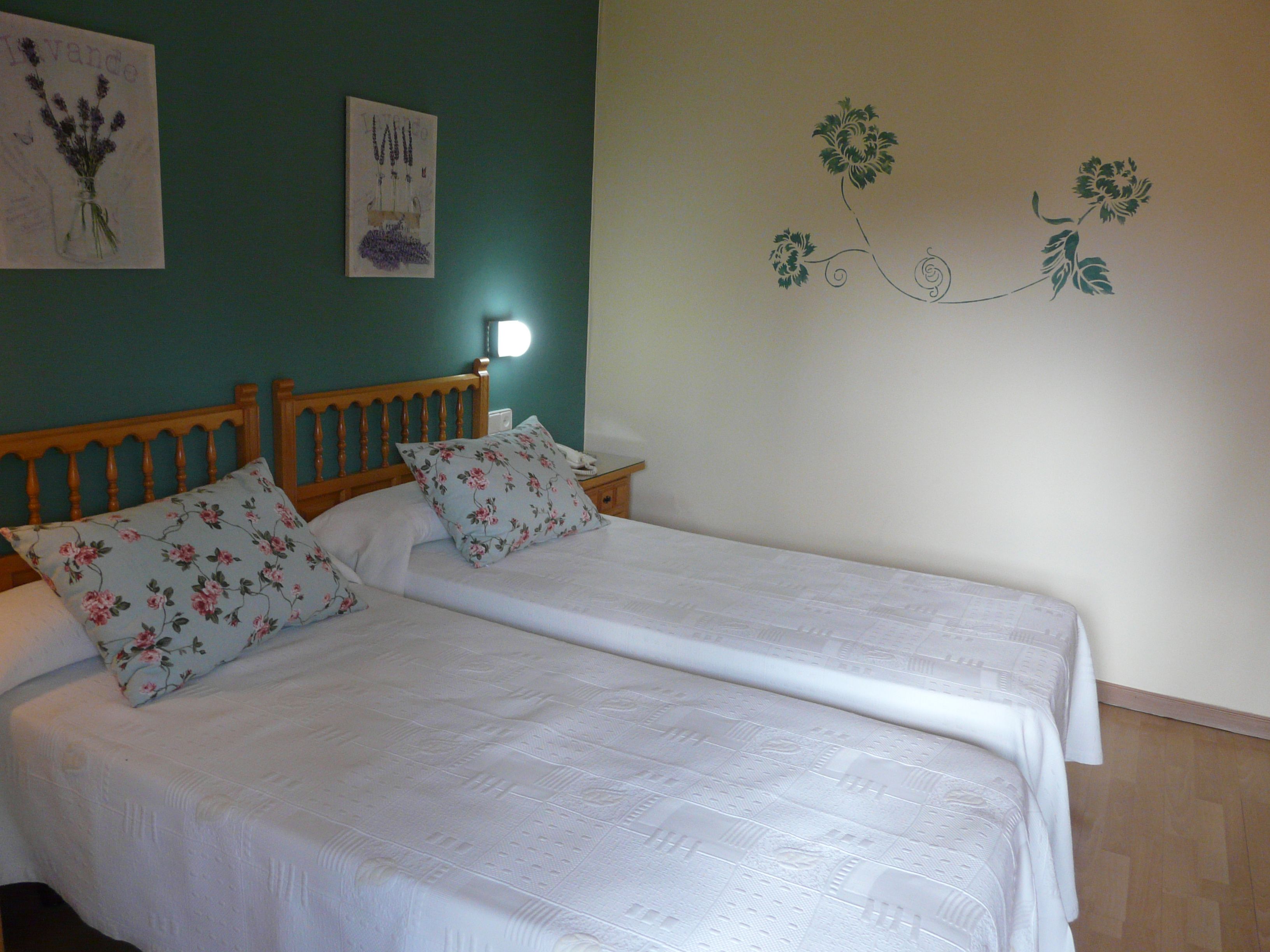A double bed with floral bedding and two pillows, a wooden headboard, a bedside table with a lamp, and a wall with a floral decal.