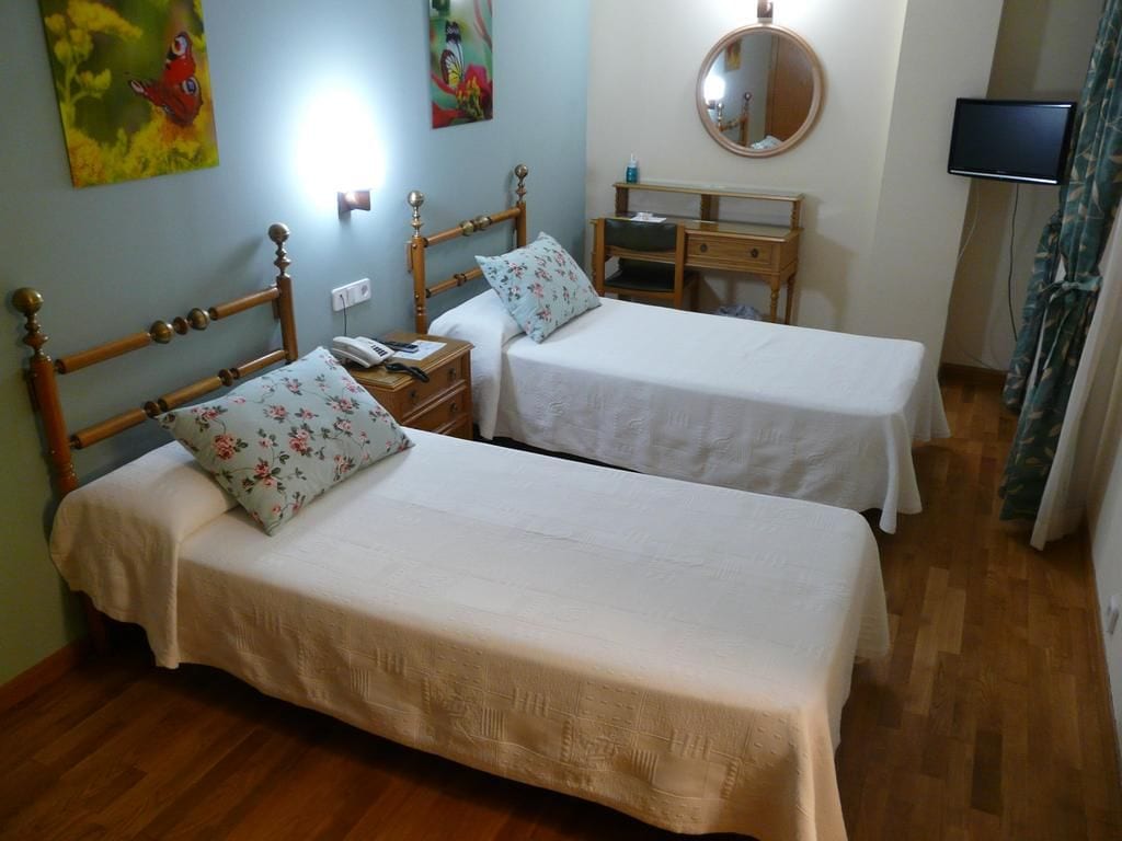 Una habitación con dos camas individuales, una mesita de noche de madera con un teléfono, un escritorio con una silla, un espejo redondo y un televisor en la pared.
