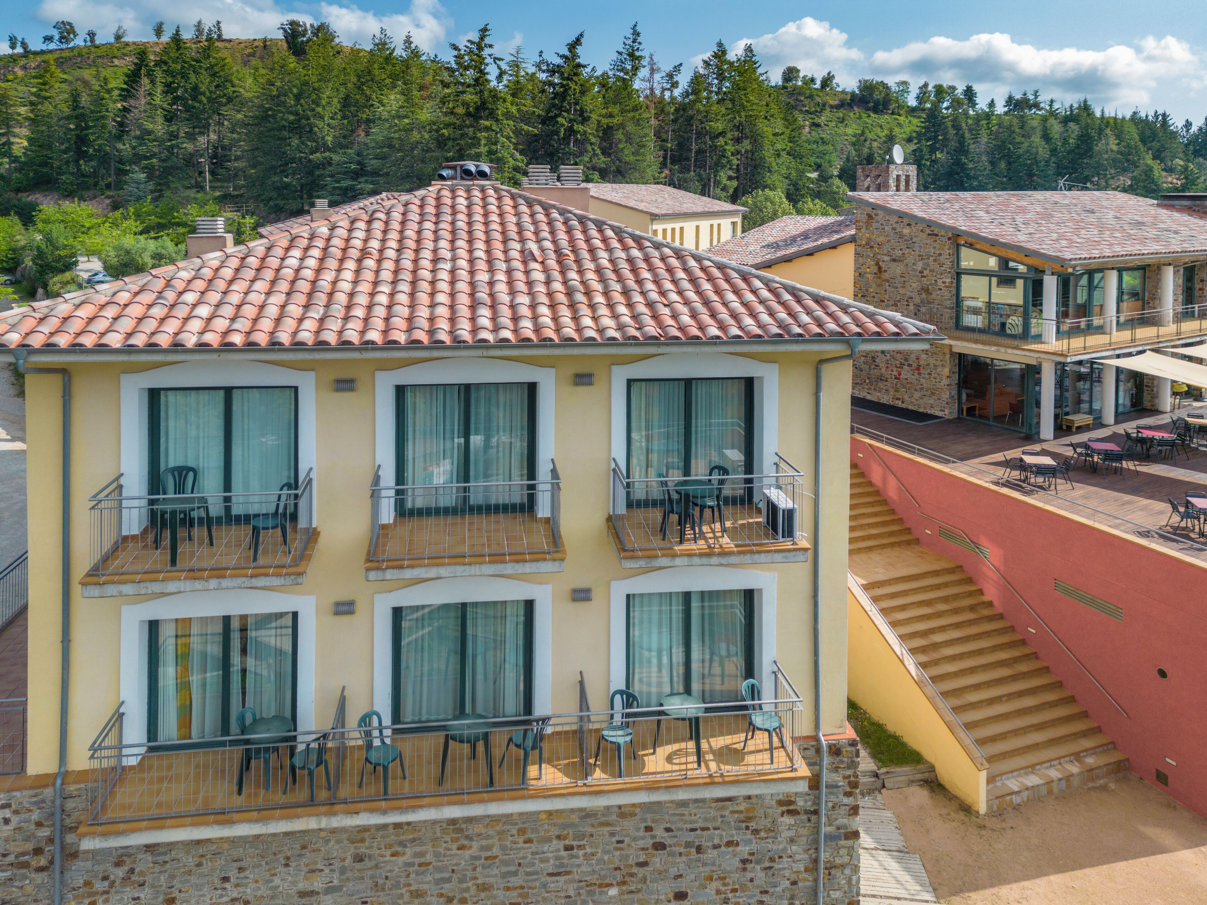 A multi-story hotel with balconies, a terrace, and a view of a forested hillside.
