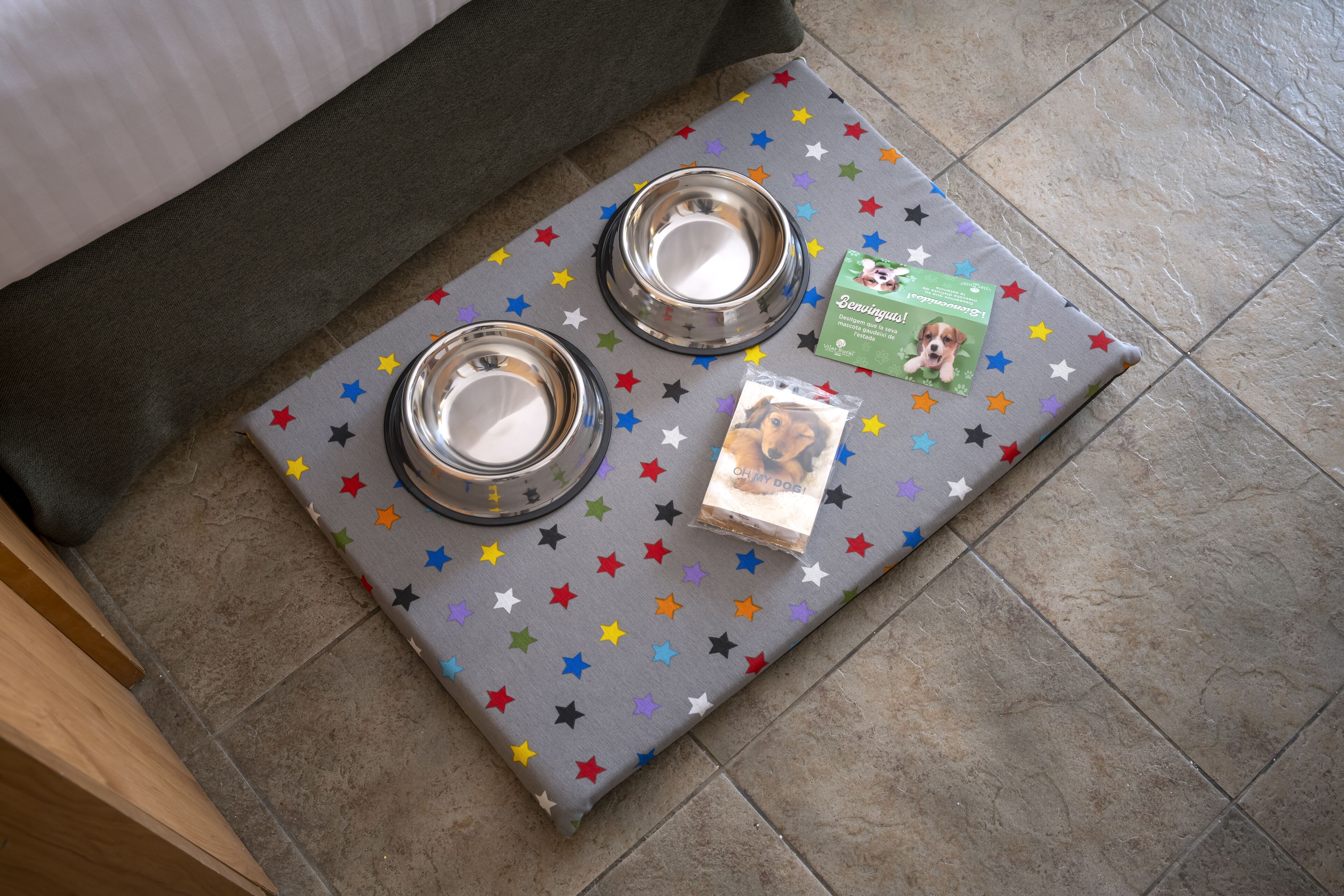 A pet bed with two metal bowls and a dog food package on a star-patterned mat.