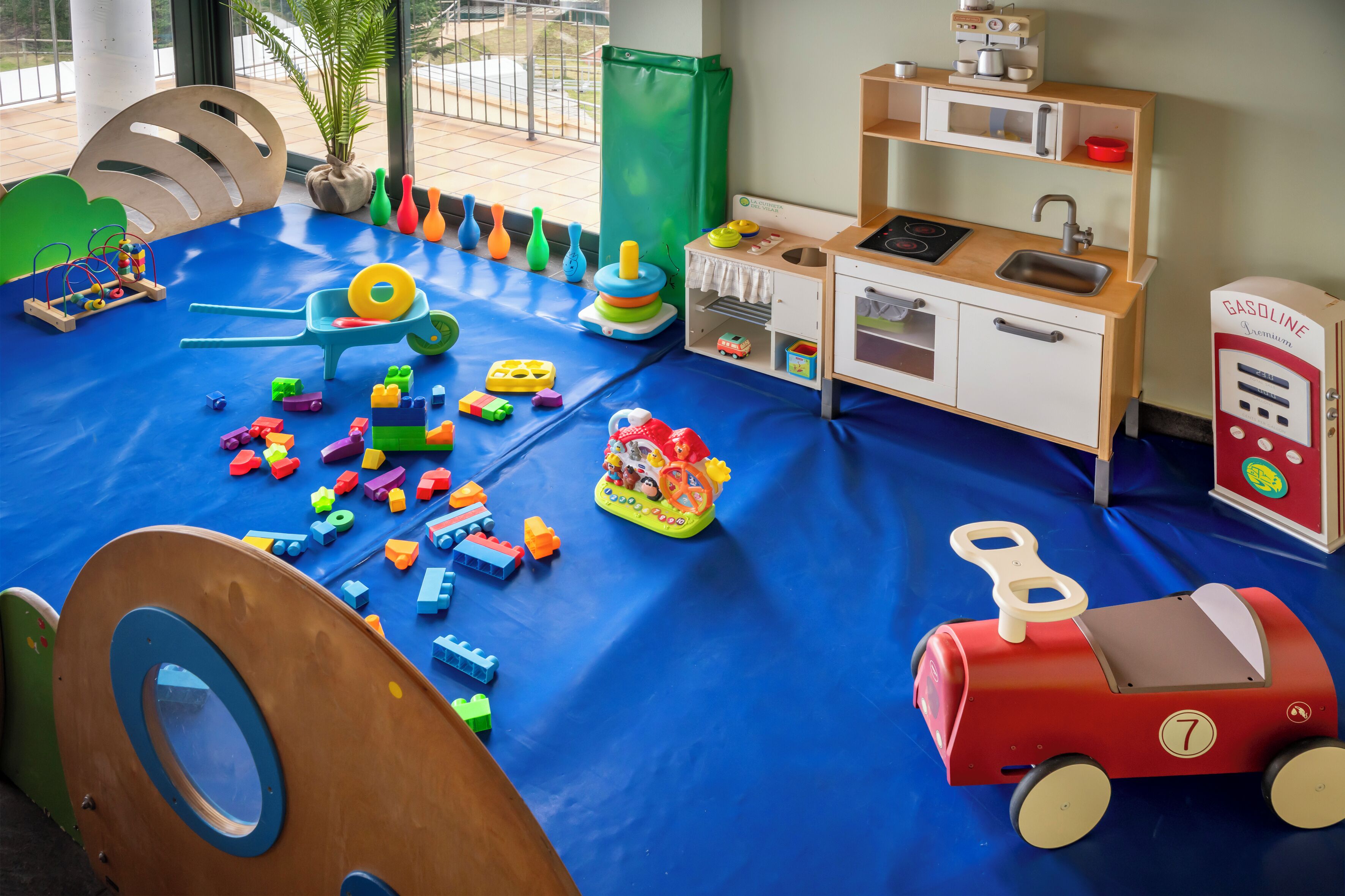 Een kinderkamer met een blauwe speelmat, een houten keukentje en diverse speelgoed.