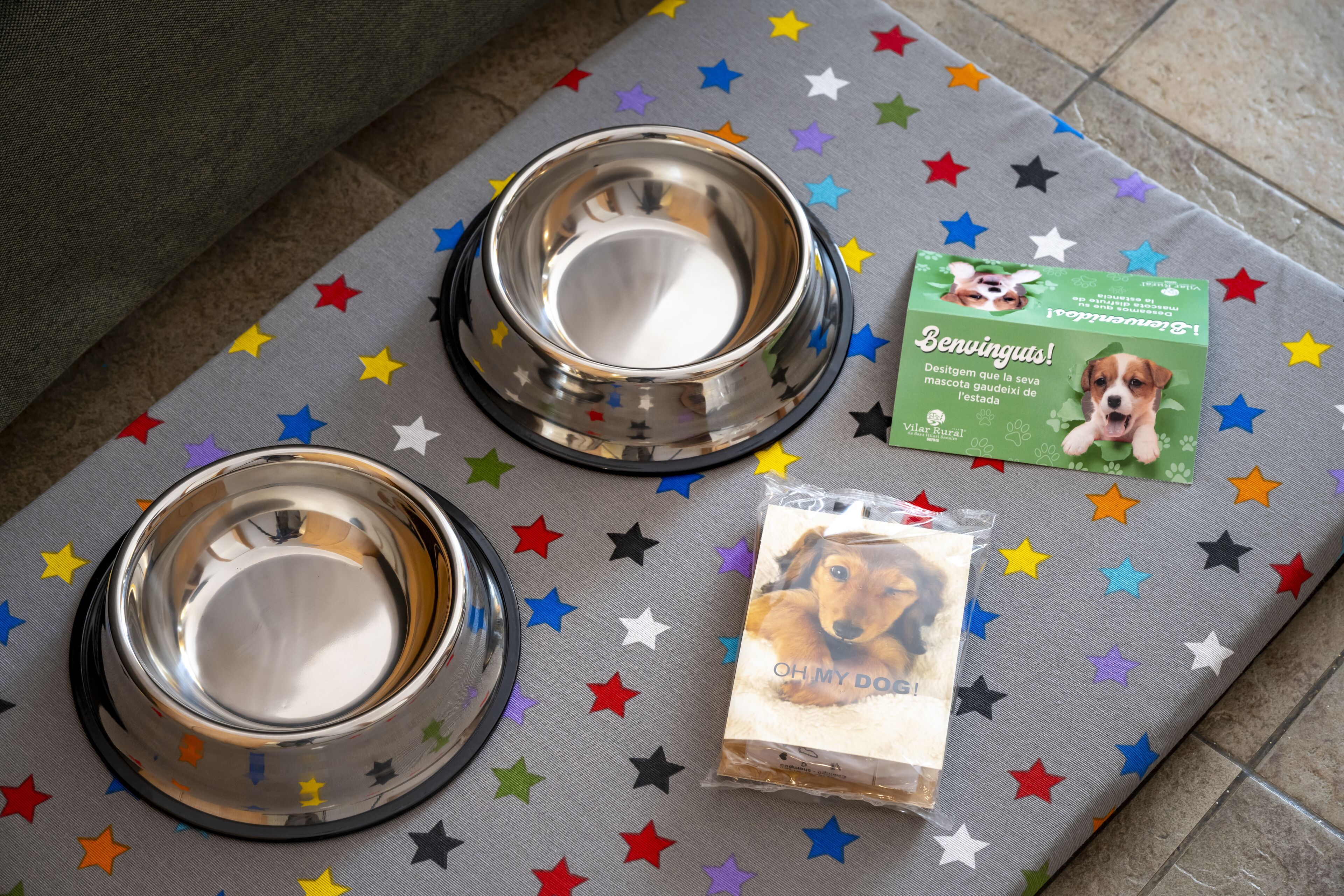 Dos tazones de acero inoxidable para mascotas sobre una superficie de tela estampada con motivos de estrellas, junto con un paquete de comida para perros y una tarjeta verde con la imagen de un perro.