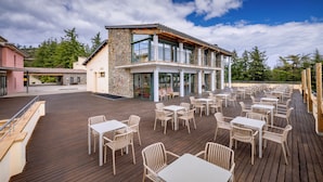 A spacious outdoor deck with multiple tables and chairs, a stone building with a balcony, and a clear view of the surrounding trees. - Vilar Rural de Sant Hilari by Serhs Hotels (Sant Hilari Sacalm)