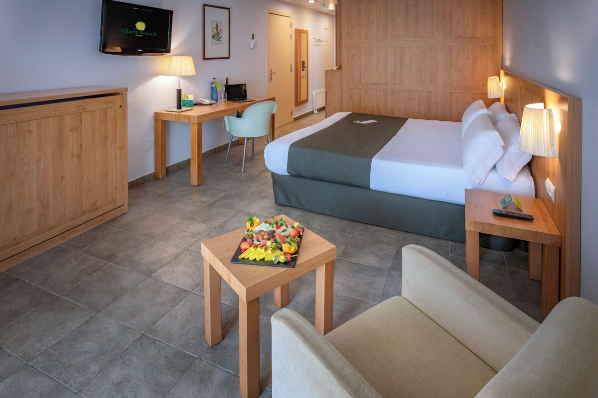 A hotel room with a bed, a desk, a chair, and a small table with fruit.