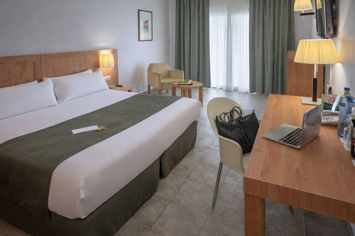 Una habitación de hotel con una cama grande, un escritorio con una computadora portátil, una silla, una pequeña mesa con una lámpara y un televisor en la pared.