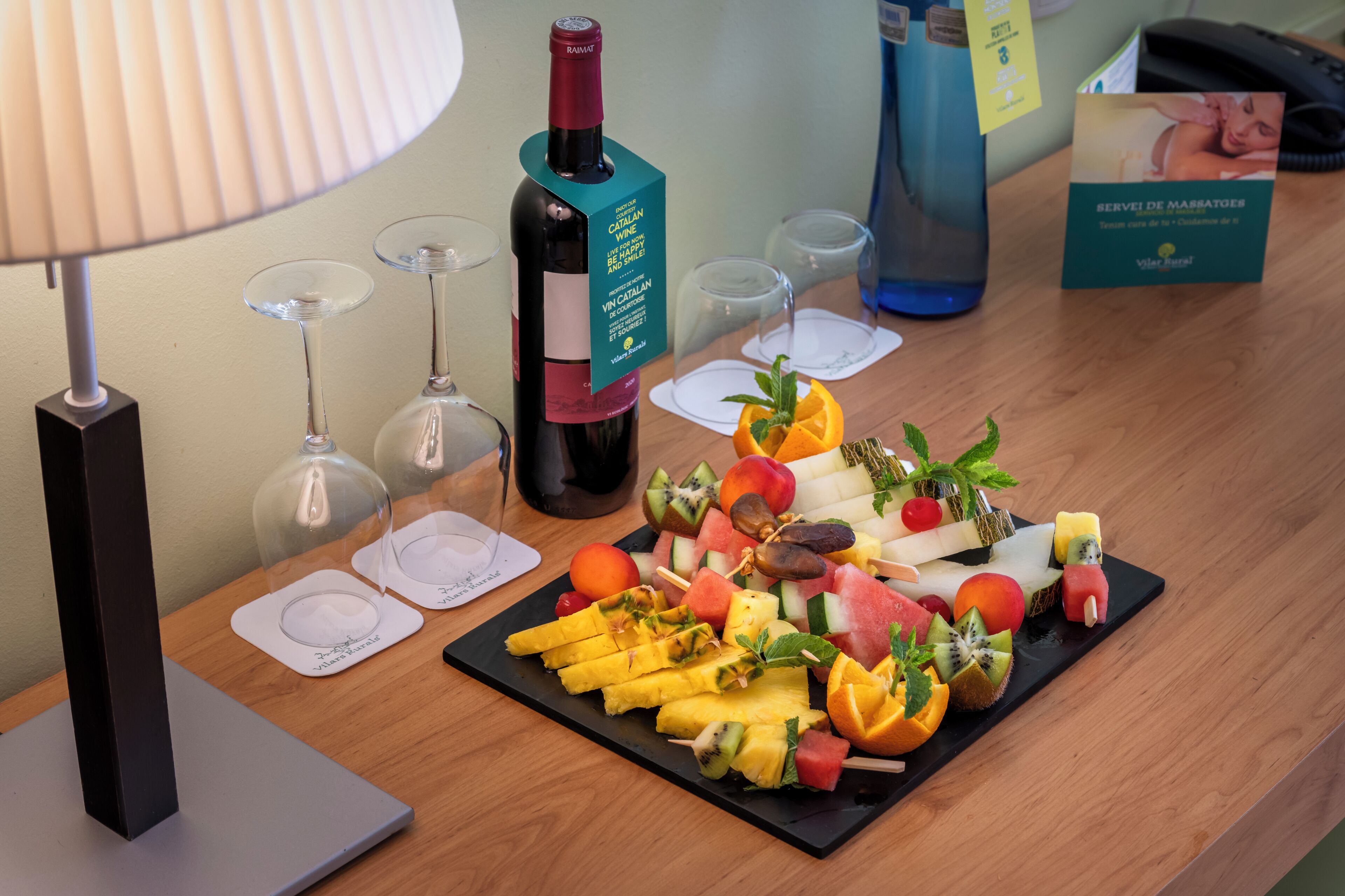 Een houten tafel met een fruitschaal, een fles wijn, glazen en een lamp.
