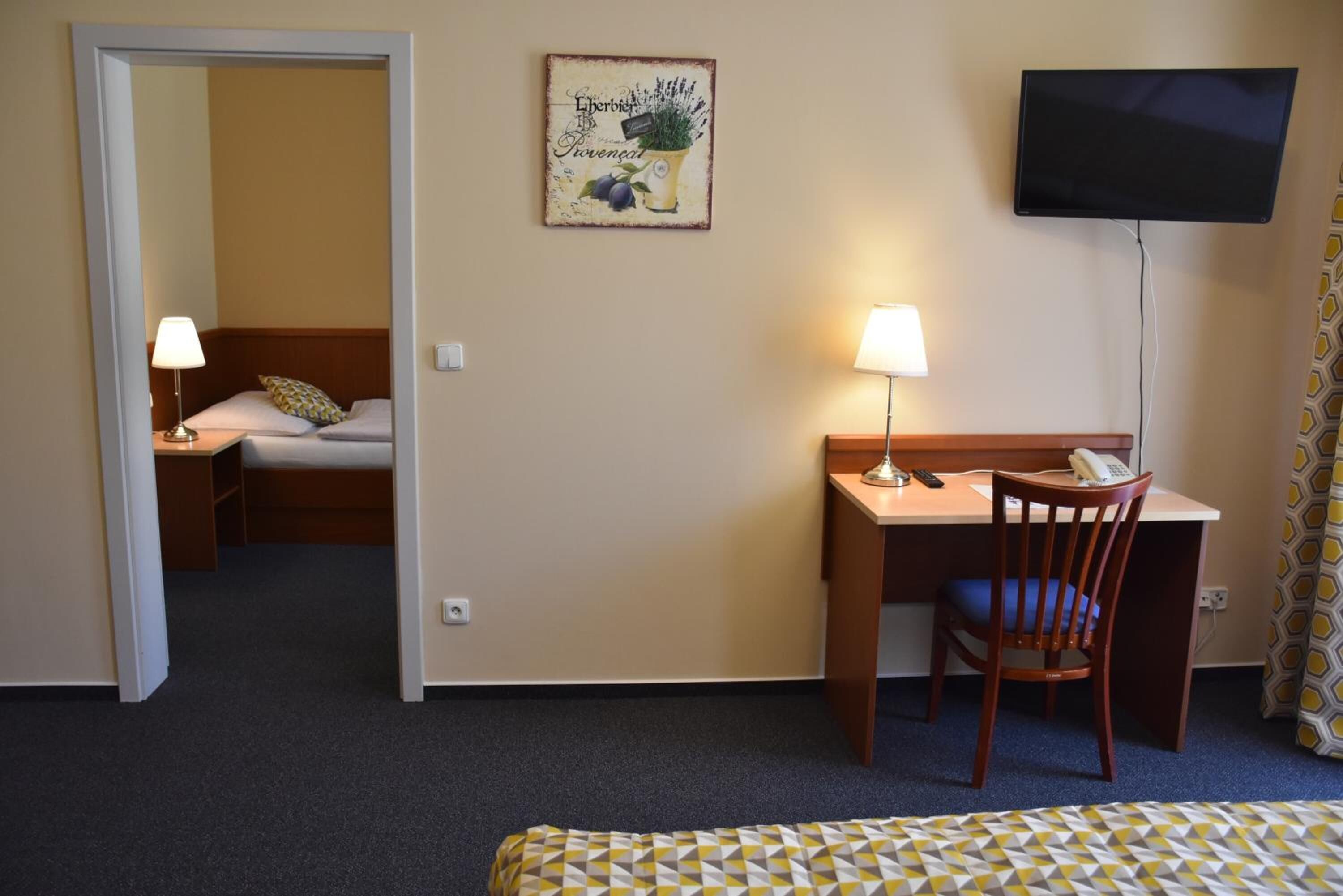 Quarto de hotel com cama, escrivaninha com cadeira, televisão e quadro na parede.