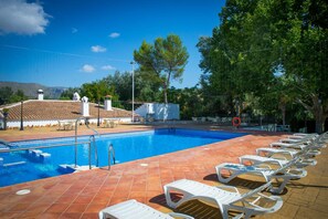Une piscine entourée de chaises longues blanches, une terrasse carrelée et une vue sur un bâtiment dont le toit est également recouvert de tuiles.