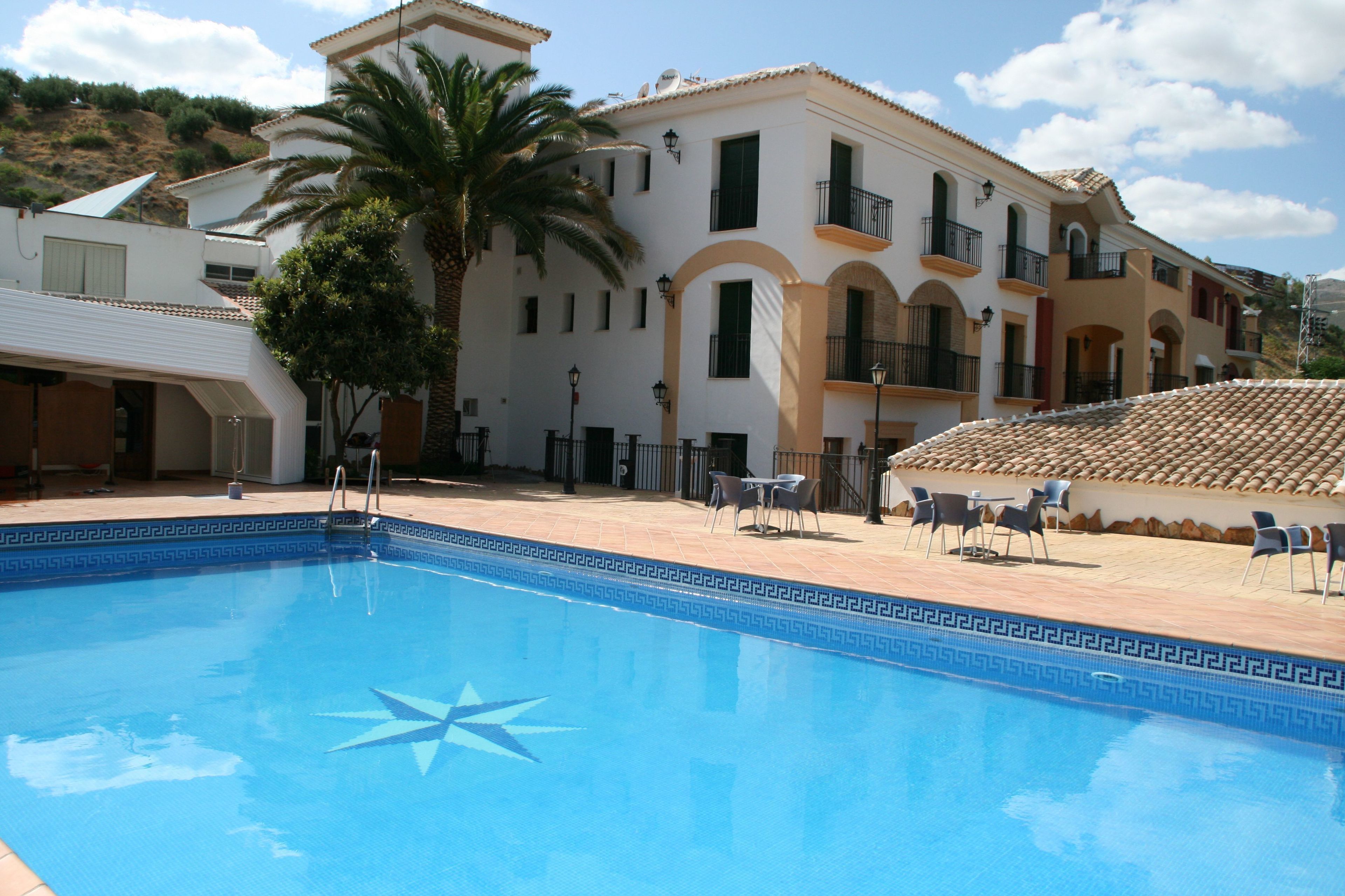 A swimming pool with a star design, surrounded by outdoor seating and a building with balconies and a tiled roof.