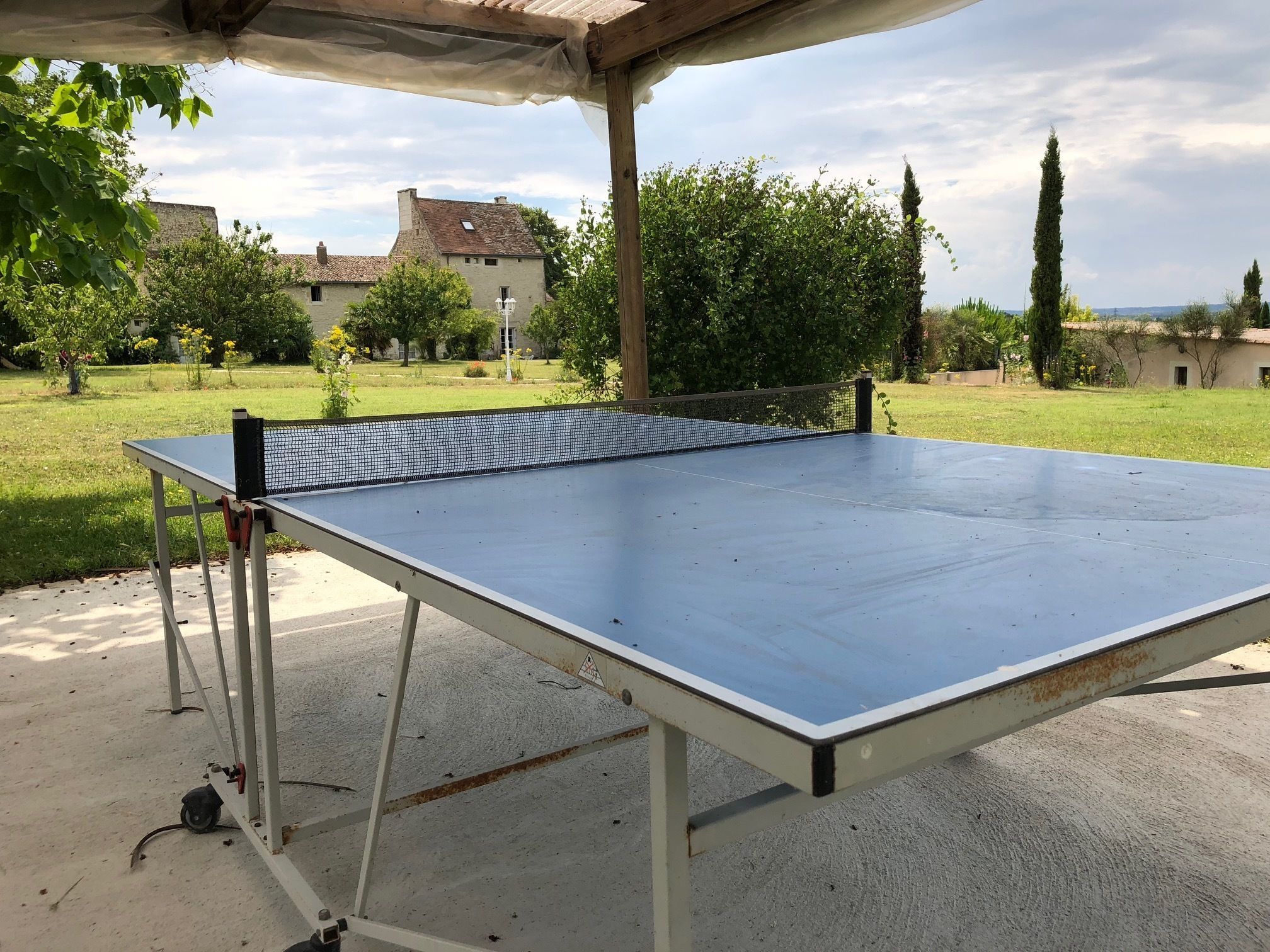 A ping pong table set up outdoors with a pergola overhead, surrounded by greenery and buildings in the background.