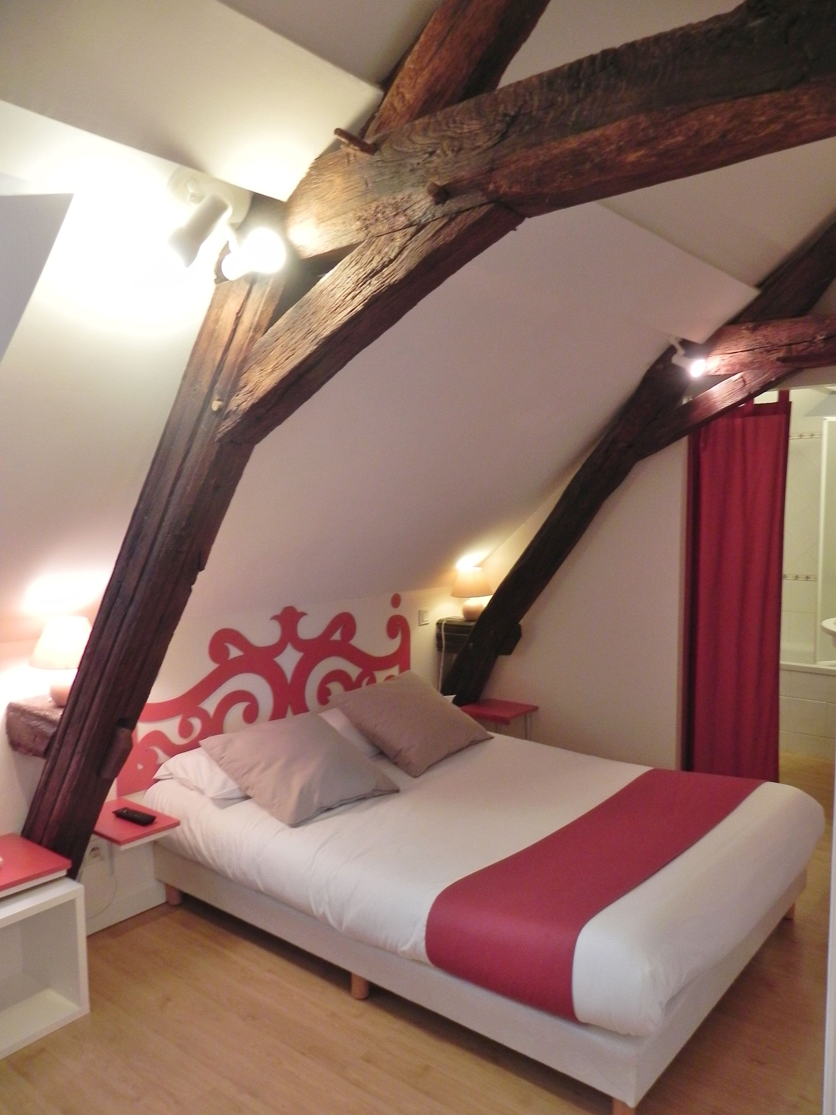 A bedroom with a sloped ceiling, a bed with white and red bedding, a red headboard, and a wooden beam structure.