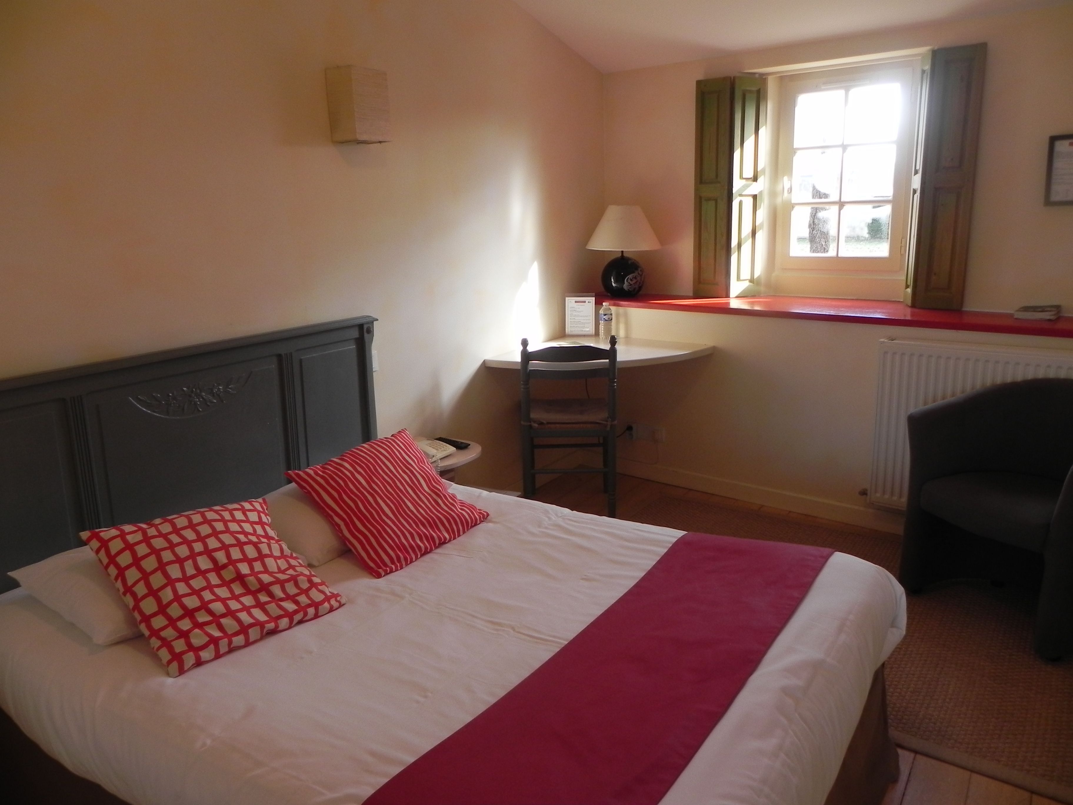 A hotel room with a bed, a desk, a chair, a lamp, and a window with shutters.