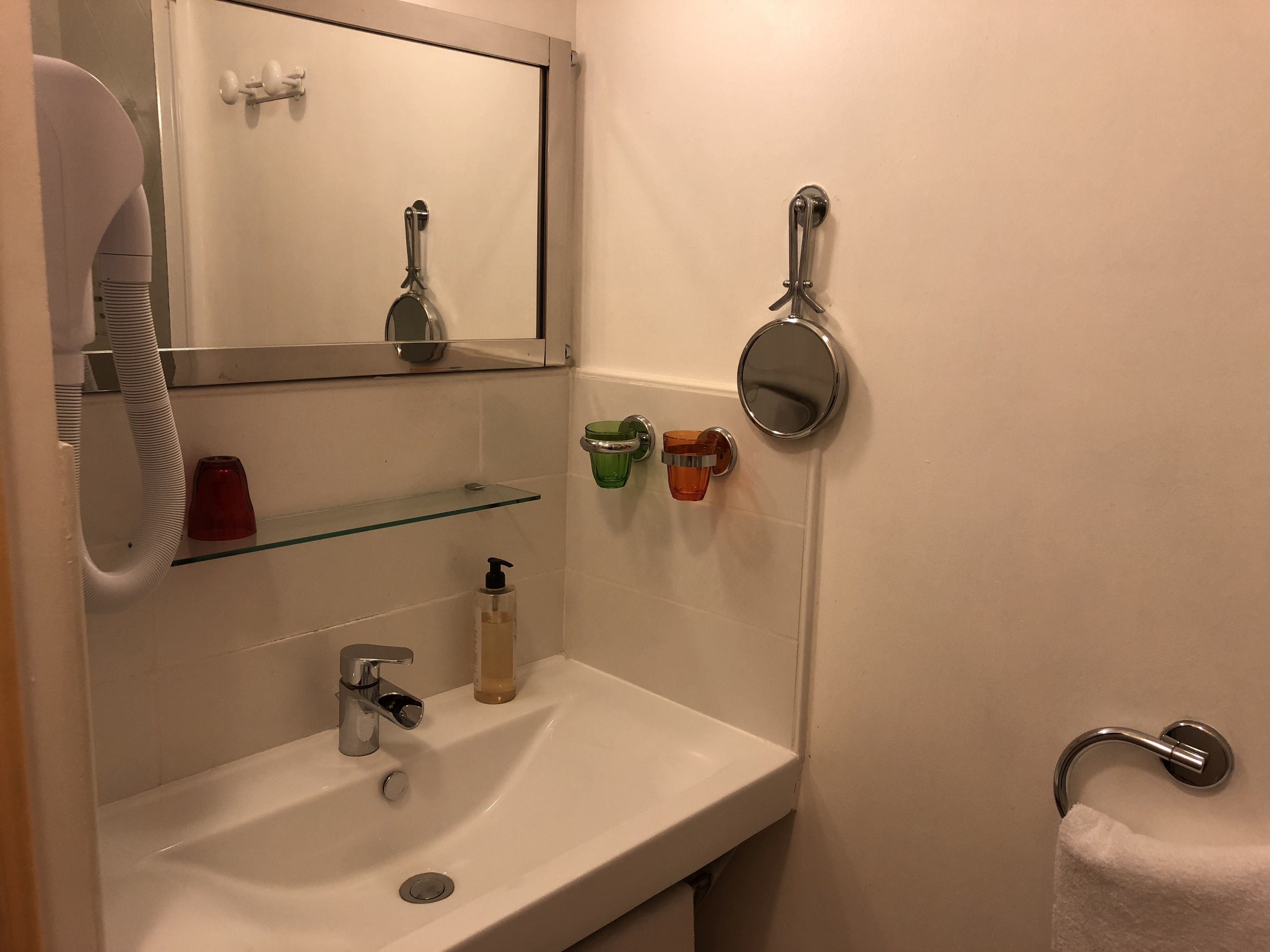 A bathroom with a white sink, a mirror, a towel rack, and a glass shelf with toiletries.