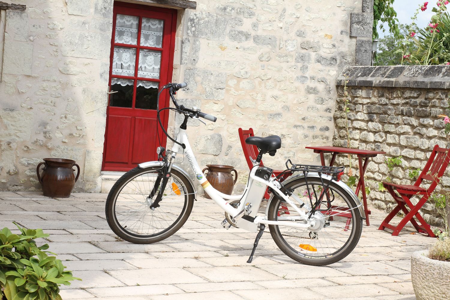 Ein weißes Fahrrad steht vor einem Steinhaus mit roter Tür und Fenstern.