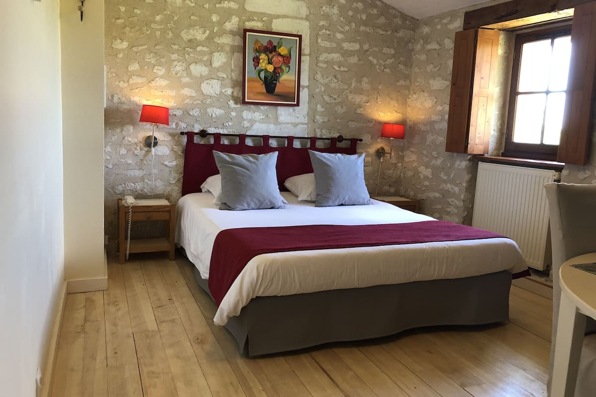 Una cama con cabecera, dos mesitas de noche, una lámpara roja, un cuadro y una ventana con cortinas.