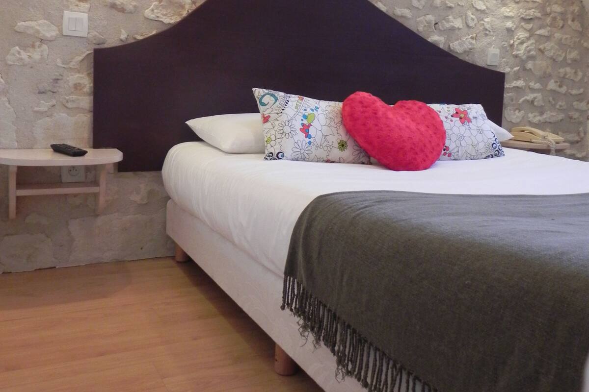 Un dormitorio con pared de piedra, una mesita de noche de madera, una cama con ropa blanca, un cabecero oscuro y una almohada roja con forma de corazón.
