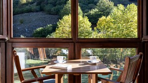 A wooden table with two white cups and saucers, set against a backdrop of a mountainous landscape through large windows.