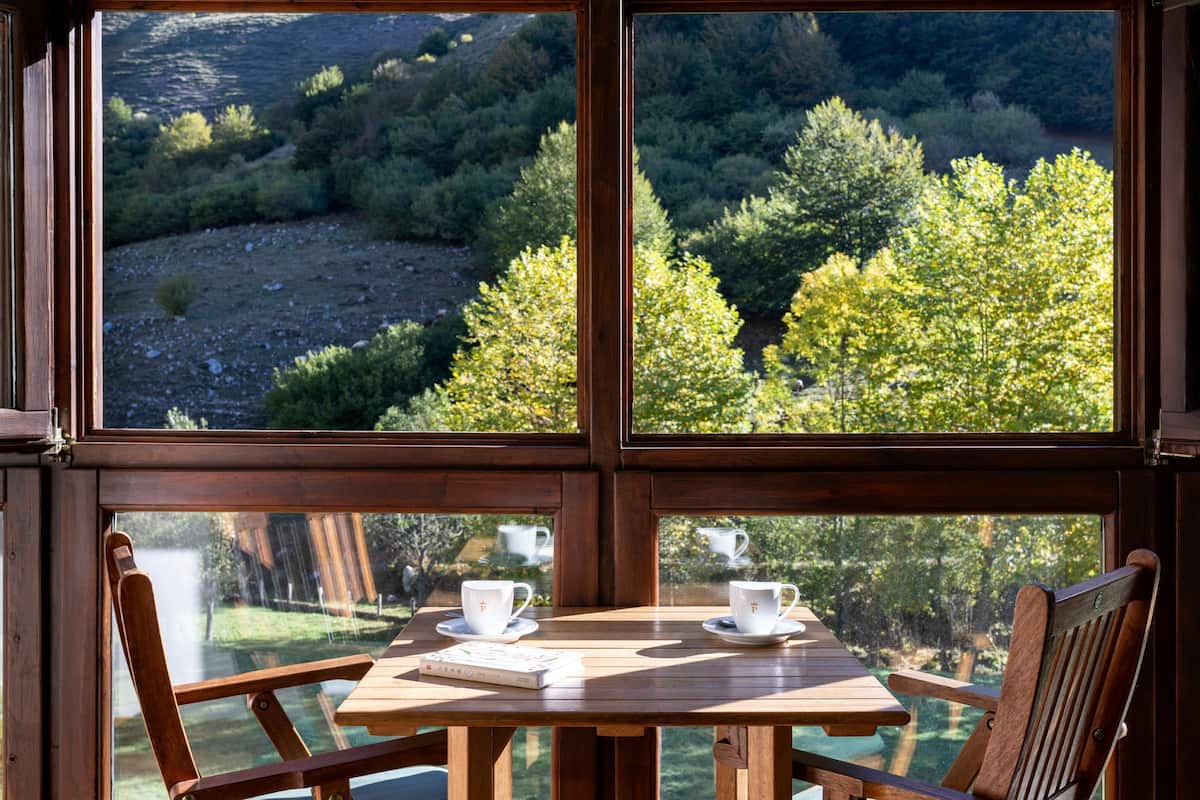 Una mesa de madera con dos tazas y platillos blancos, frente a un paisaje montañoso visible a través de grandes ventanales.