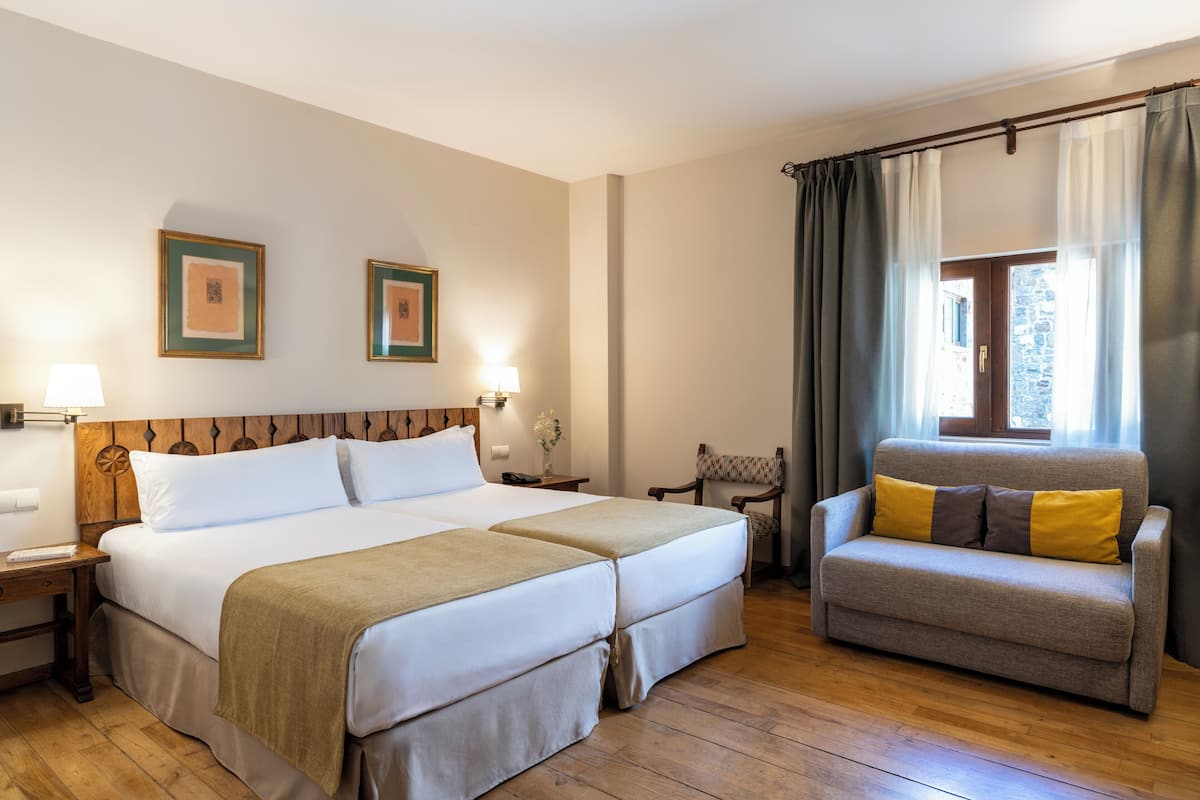 Habitación de hotel con cama doble, sofá, sillón, mesita de noche, lámpara y cuadros en la pared.