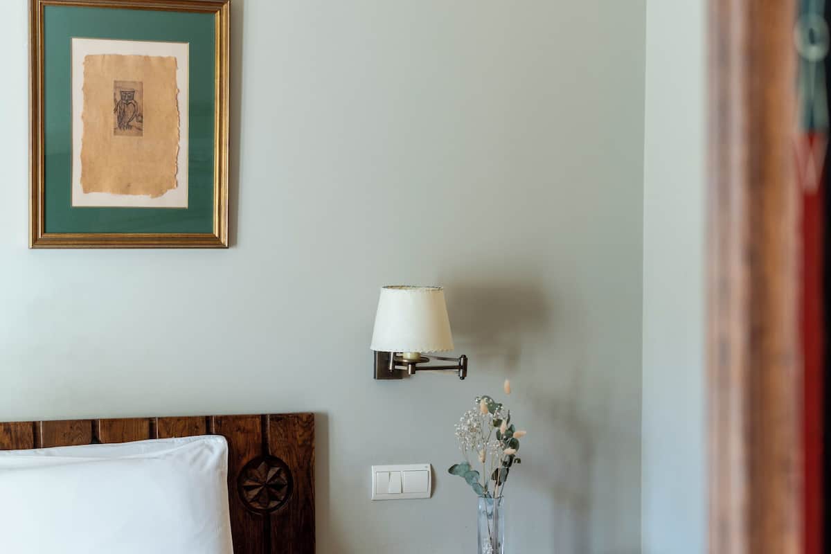 Una cama bien hecha con cabecera de madera, una mesita de noche con una lámpara, un cuadro enmarcado en la pared y un jarrón con flores sobre la mesa.