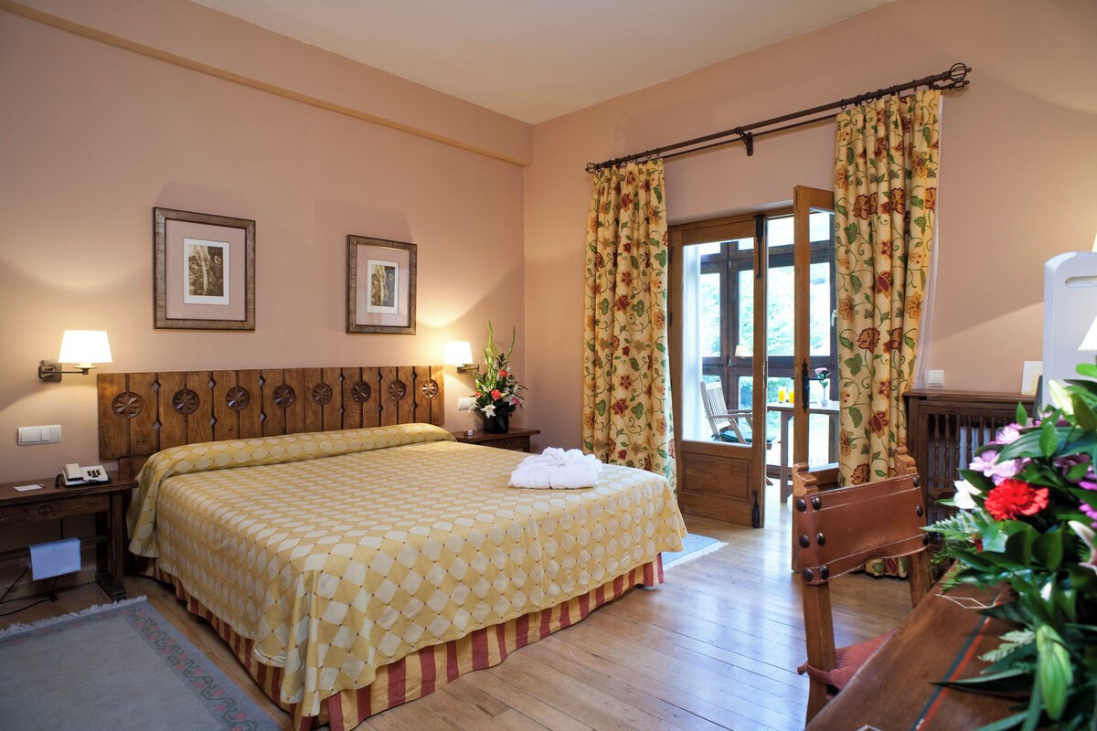 A bedroom with a large bed, wooden headboard, two framed pictures, a nightstand with a lamp, a wooden chair, and a window with curtains.