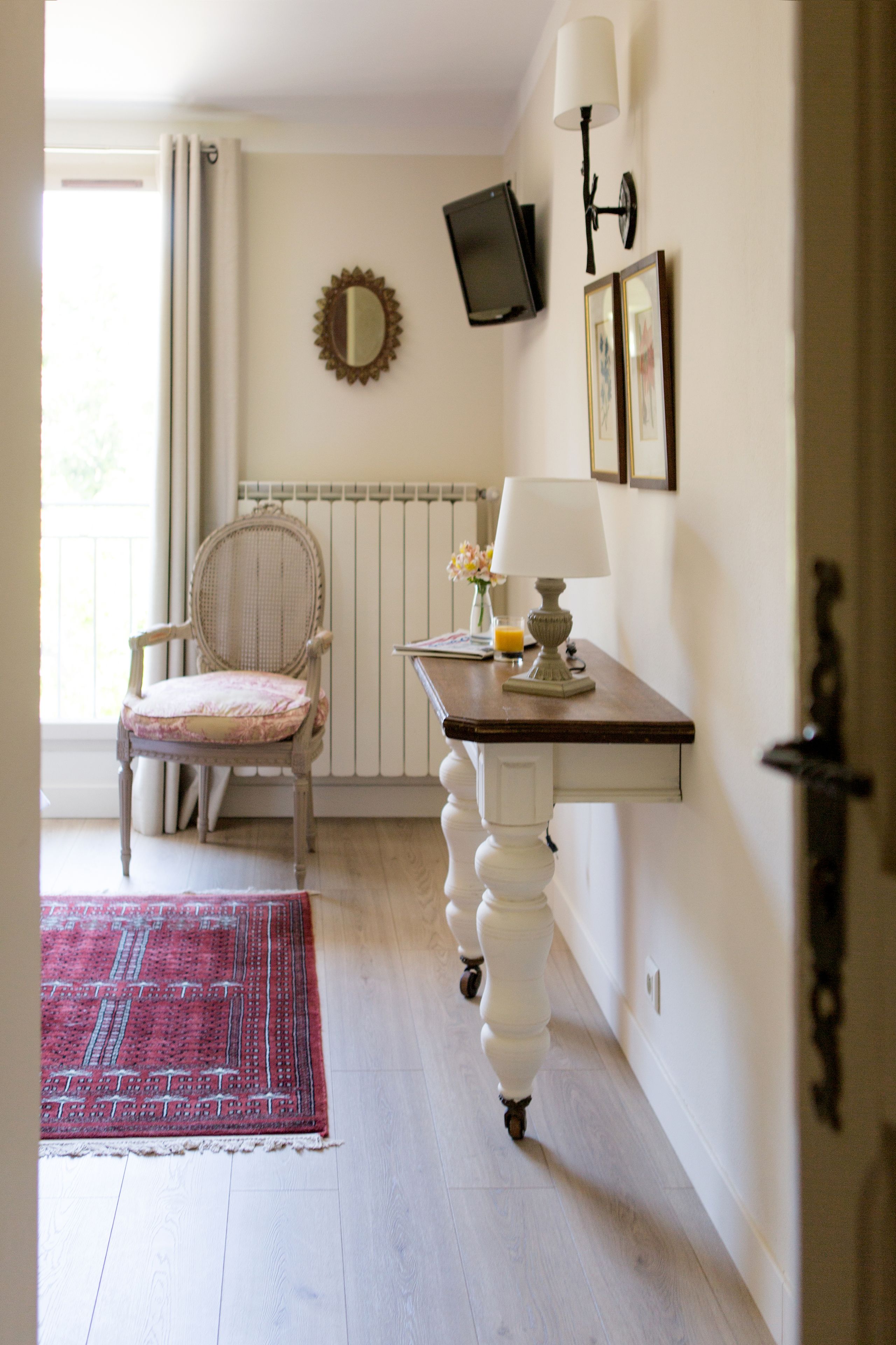 A room with a wooden floor, a red patterned rug, a chair with a cushion, a small table with a lamp, a wall-mounted TV, and a mirror.
