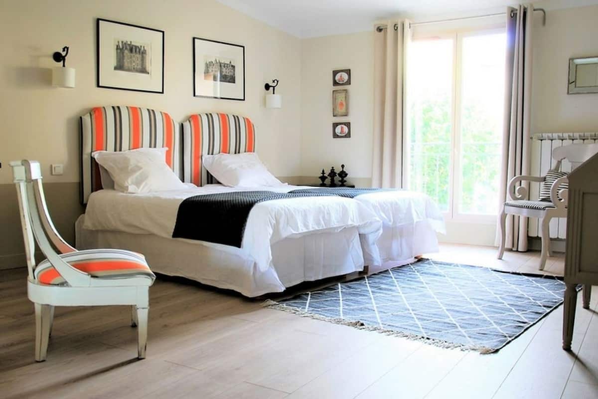 Un lit double avec des coussins à rayures, une chaise blanche, une petite table et une vue sur la verdure à l’extérieur.