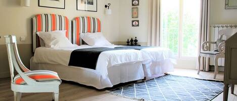 A double bed with striped pillows, a white chair, a small table, and a view of greenery outside.