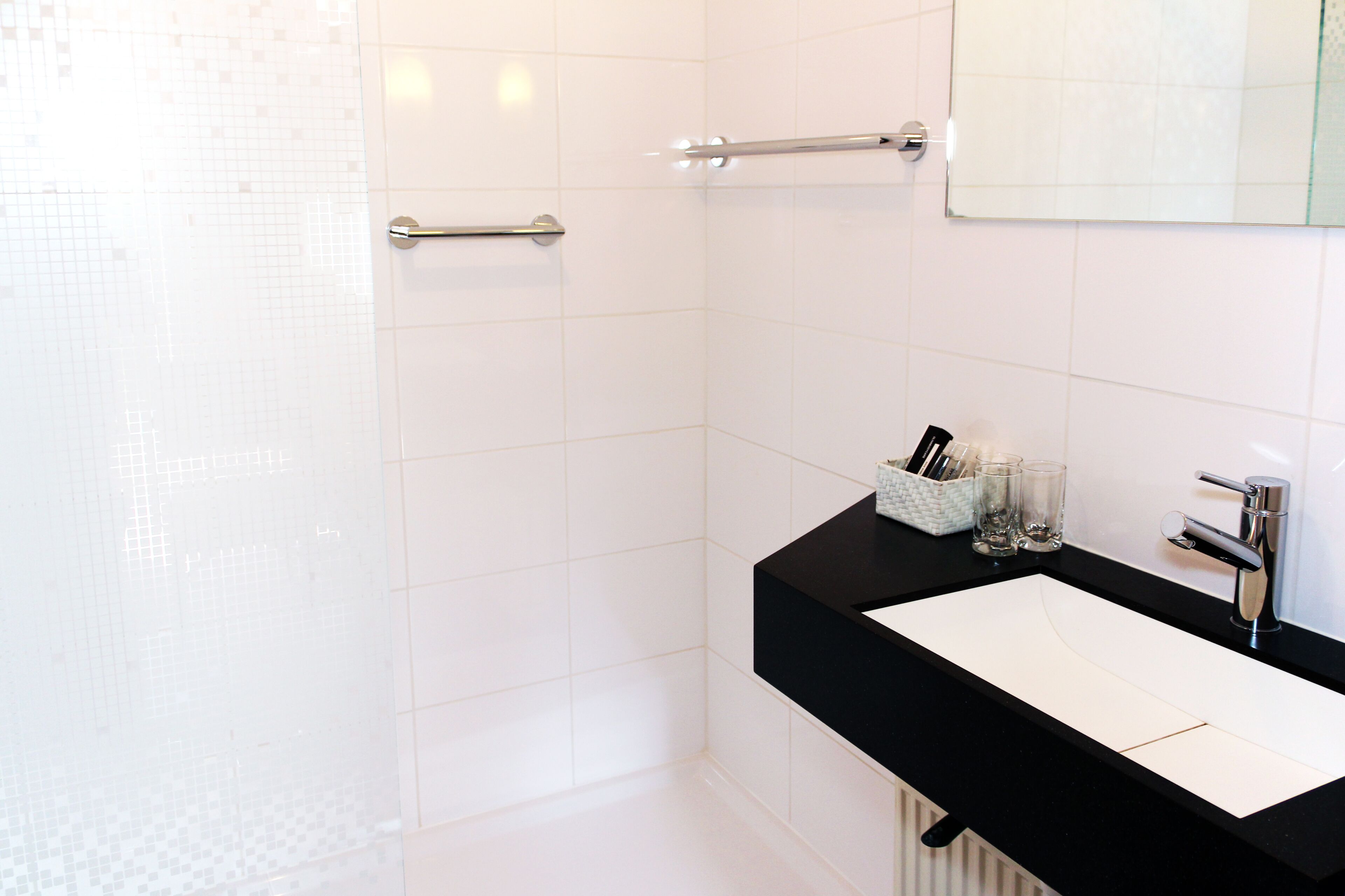 Un baño con pared de azulejos blancos, encimera negra, lavabo blanco, espejo y perchero.