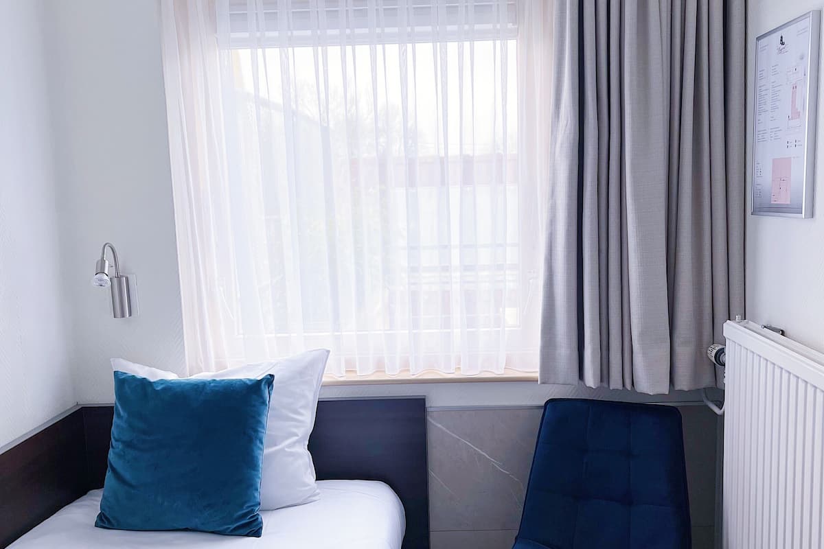 A hotel room with a bed, a chair, a window with curtains, and a radiator.