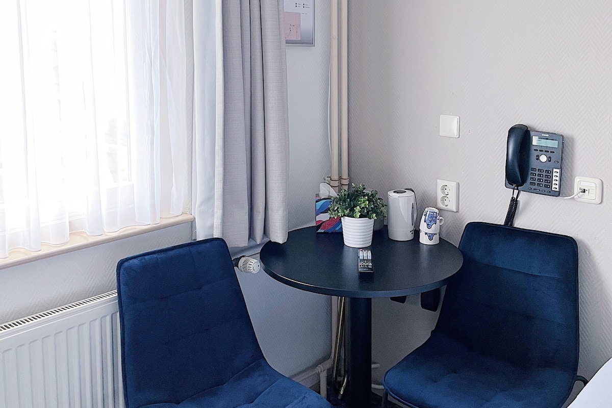 A small room with two blue chairs, a round table, a potted plant, a telephone, and a window with curtains.