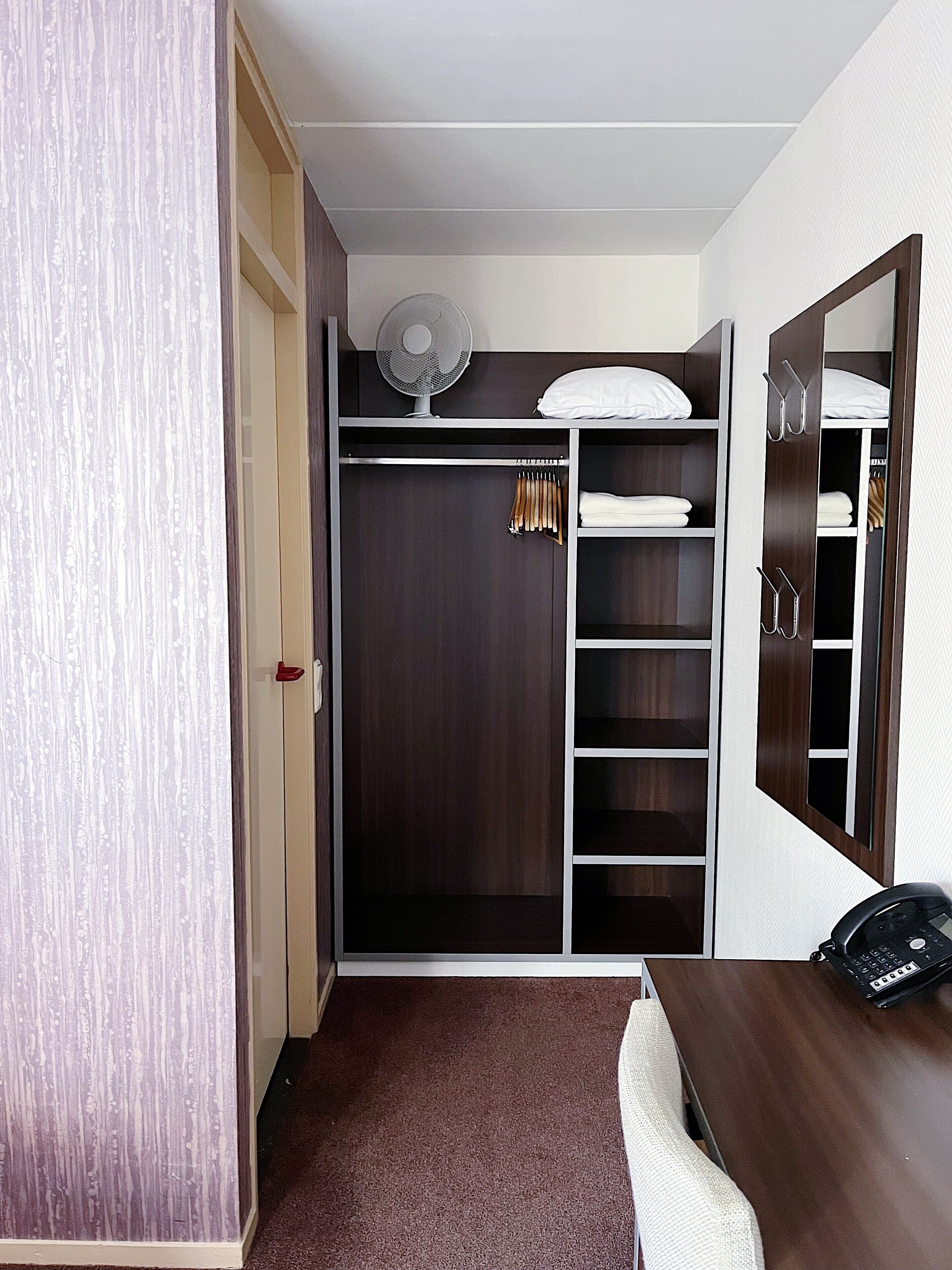 A bedroom with a dark wood wardrobe, a white bed, a desk with a laptop, and a standing fan.