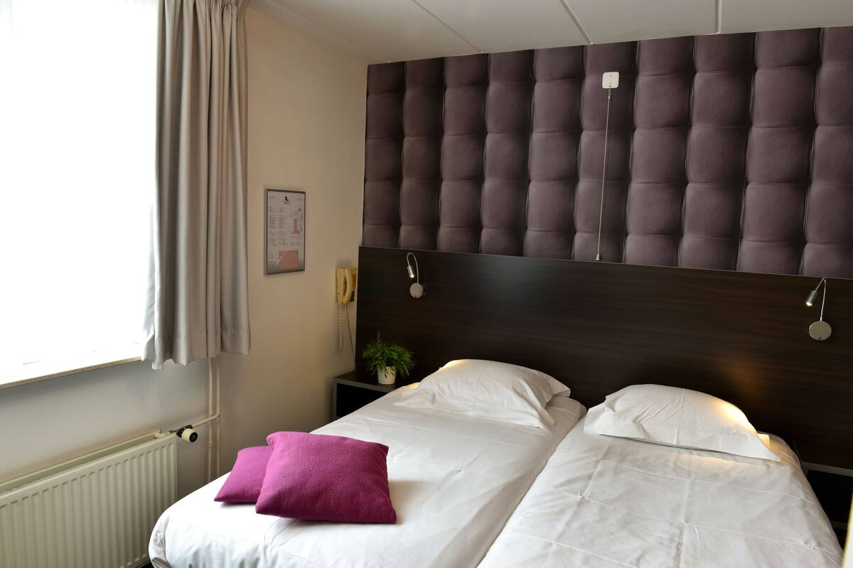 A hotel room with a large bed, a headboard, a bedside table with a lamp, and a window with curtains.