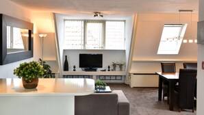 A modern living room with a flat-screen TV, a dining area with a table and chairs, and a large window with blinds. - De Magneet (Hoorn)