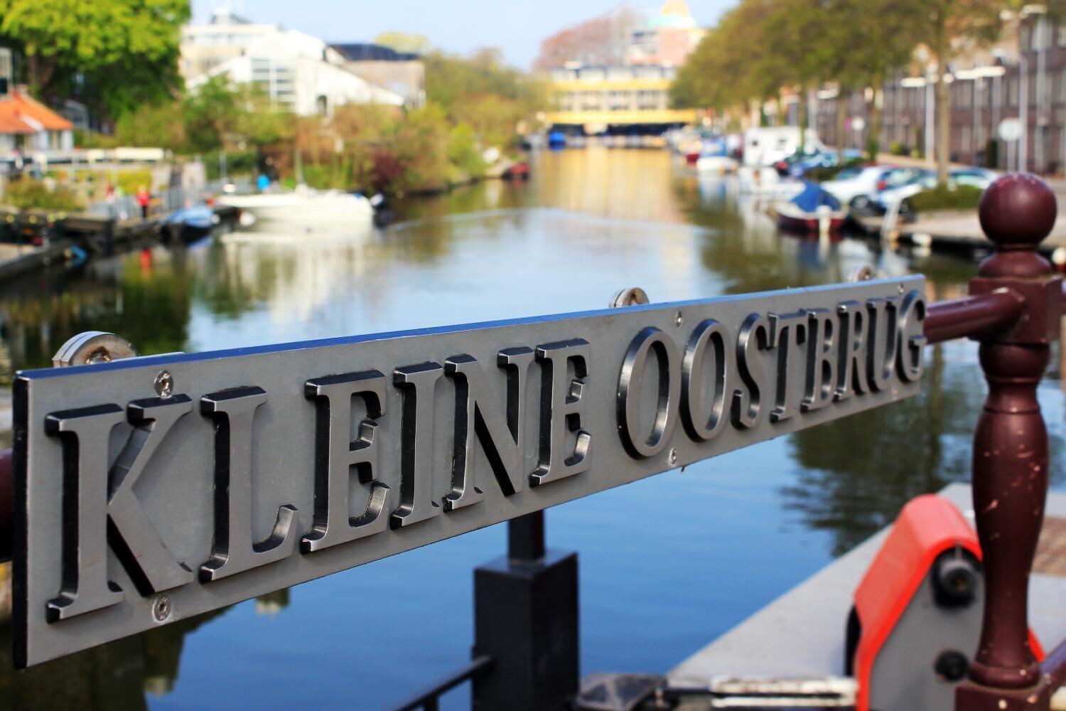 Un cartel con la inscripción 'Kleine Oosterkerk' frente a un canal con barcos y edificios al fondo.