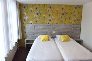 Habitación de hotel con dos camas individuales, cabecero de madera, una mesita de noche pequeña y una ventana con cortinas blancas.
