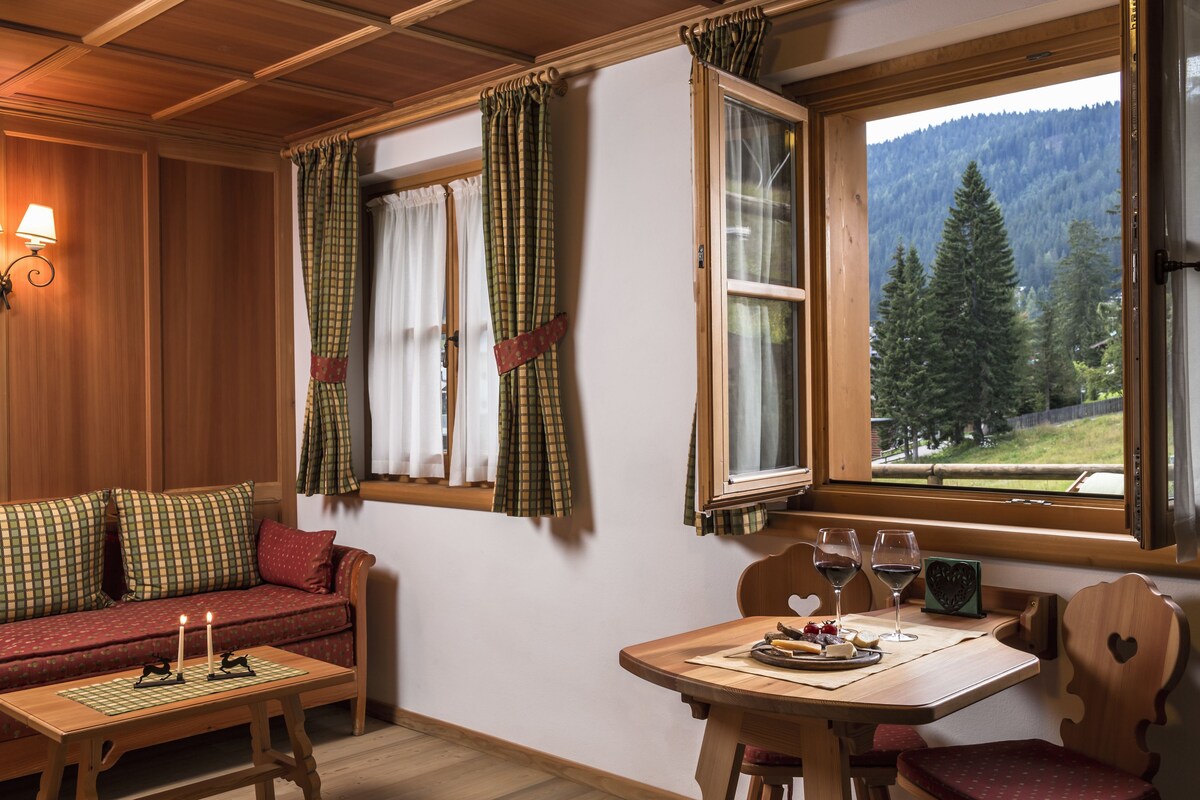 A cozy cabin interior with a wooden ceiling, a sofa with plaid cushions, a dining table with a candle, and a window with curtains overlooking a forested mountain.