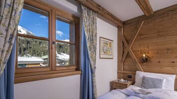 Quarto com teto de madeira, cama com roupa de cama branca, mesa de cabeceira com abajur e janela com cortinas.