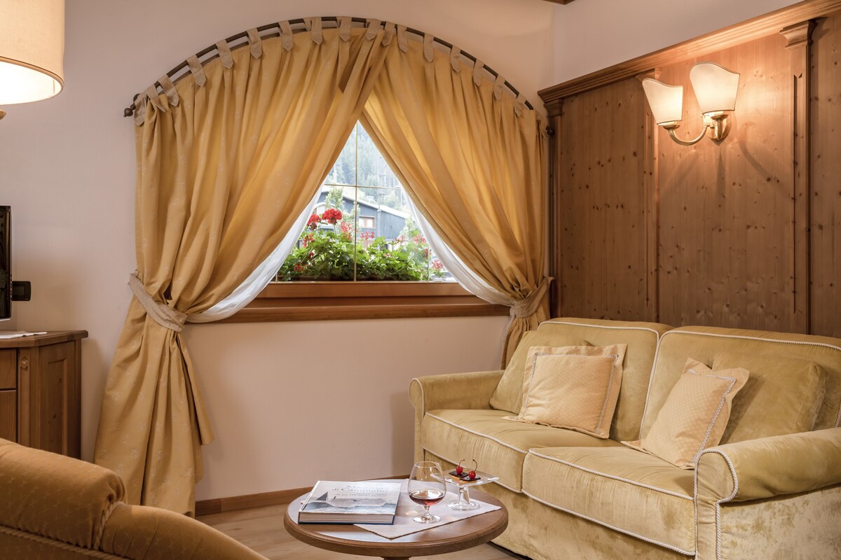 A cozy living room with a beige sofa, a small round table with a book and a glass of wine, and a window with curtains.