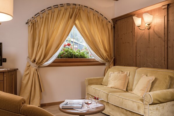 A cozy living room with a beige sofa, a small round table with a book and a glass of wine, and a window with curtains. - Hotel Chalet del Sogno (Pinzolo)