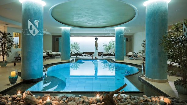 A pool area with a person walking, surrounded by columns and lounge chairs. - Hotel Chalet del Sogno (Pinzolo)