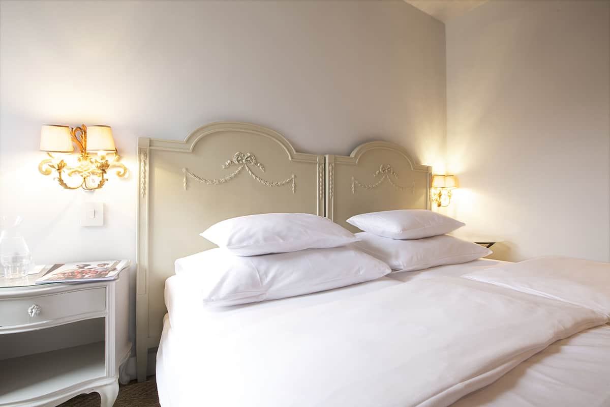 A neatly made bed with white linens and pillows, a bedside table with a lamp, and a wall-mounted light fixture.