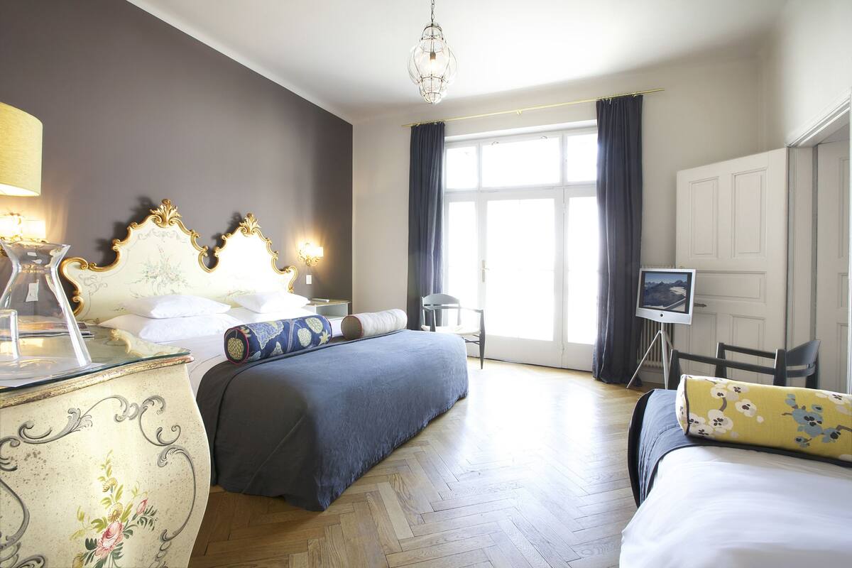 A hotel room with a large bed, a smaller bed, a desk with a lamp, a chair, a painting on the wall, and a view of the outdoors.