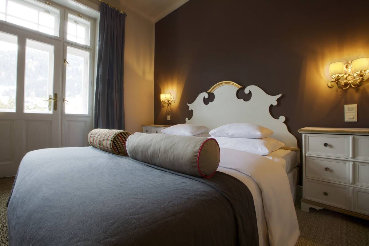 A neatly made bed with a headboard, a bedside table with a lamp, and a window with curtains.