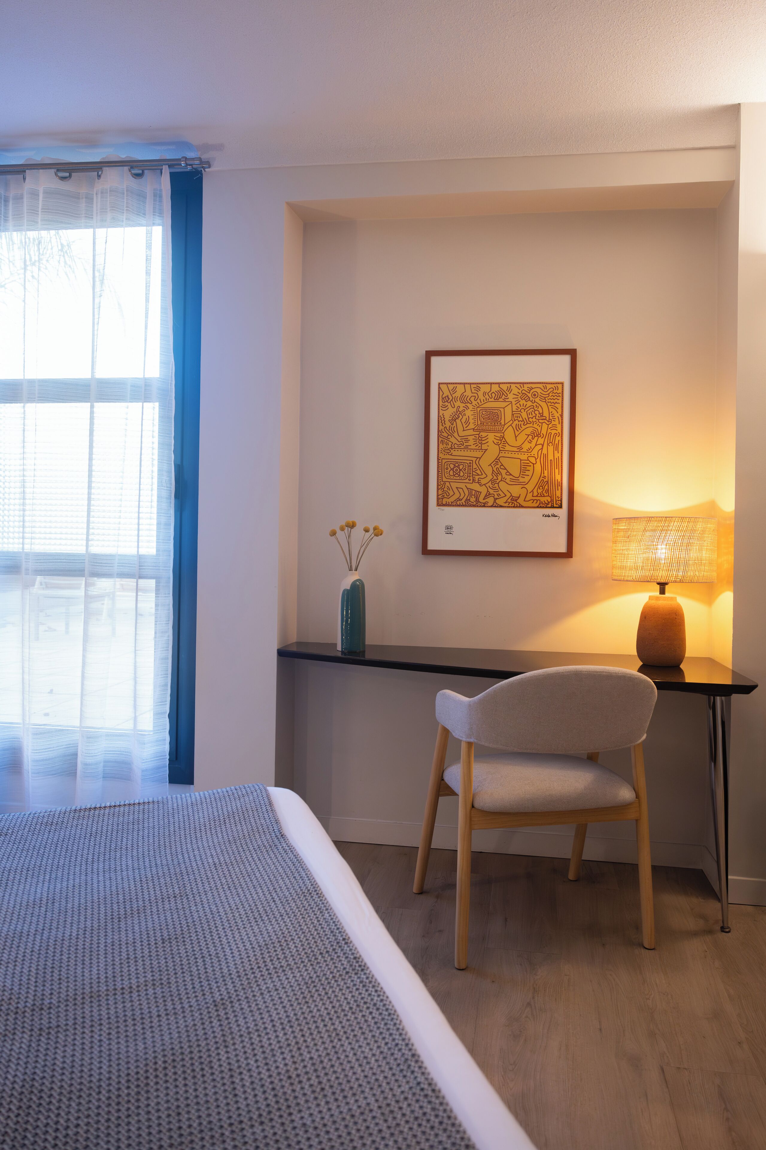 A hotel room with a bed, a desk, a chair, a lamp, and a framed artwork on the wall.