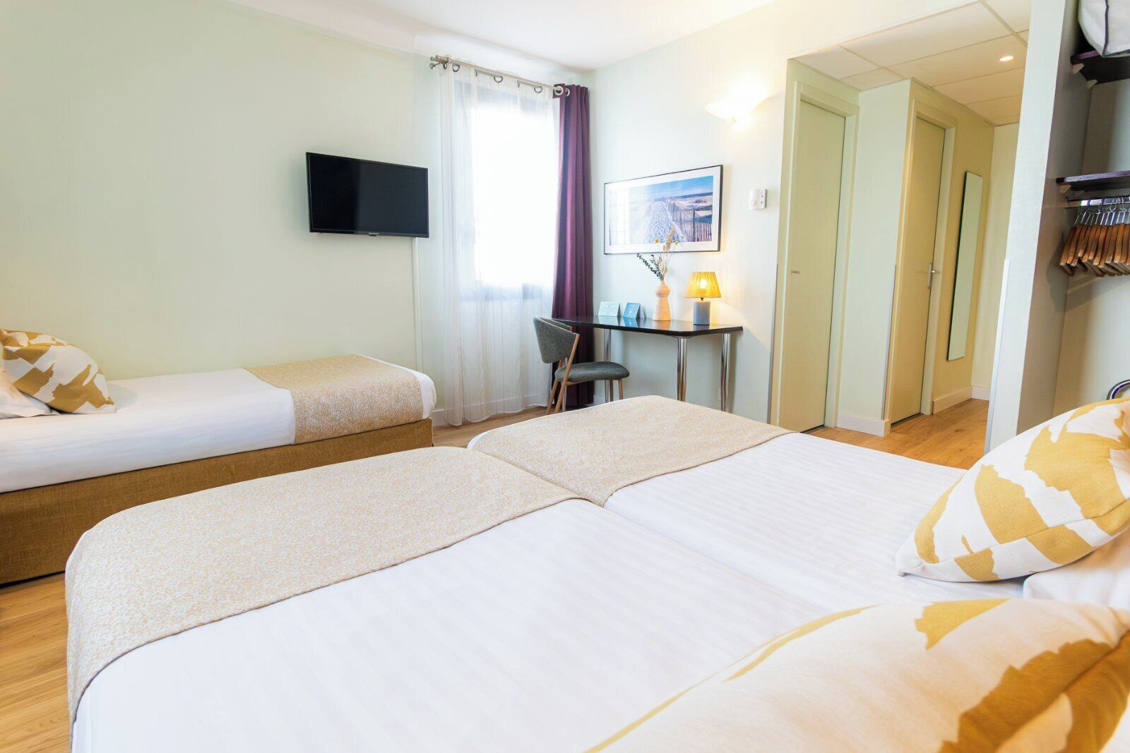 A hotel room with two single beds, a desk with a chair, a TV mounted on the wall, and a door leading to another room.