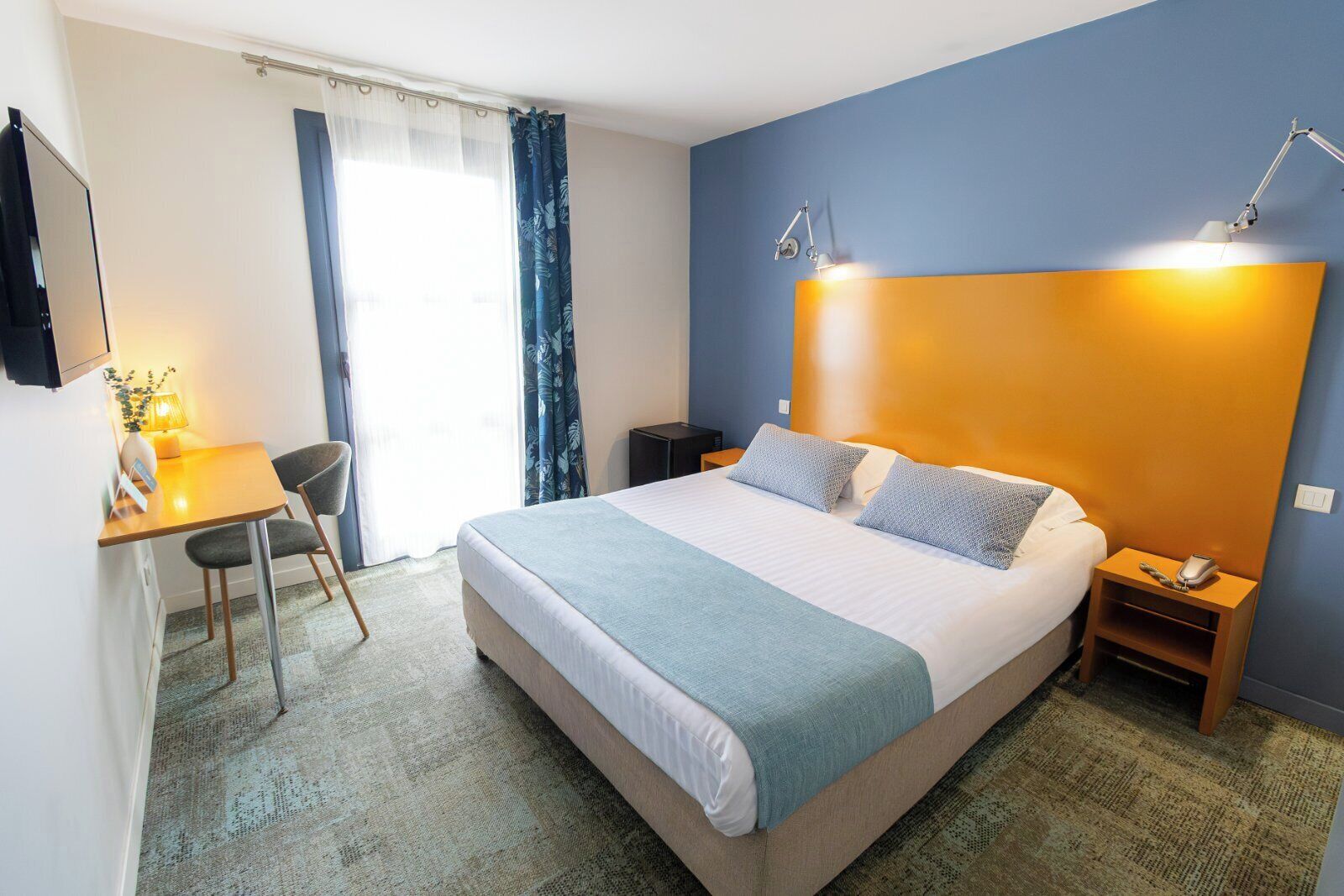 Une chambre d’hôtel comprenant un lit, des tables de chevet, un bureau avec une chaise, une lampe et une fenêtre avec des rideaux.