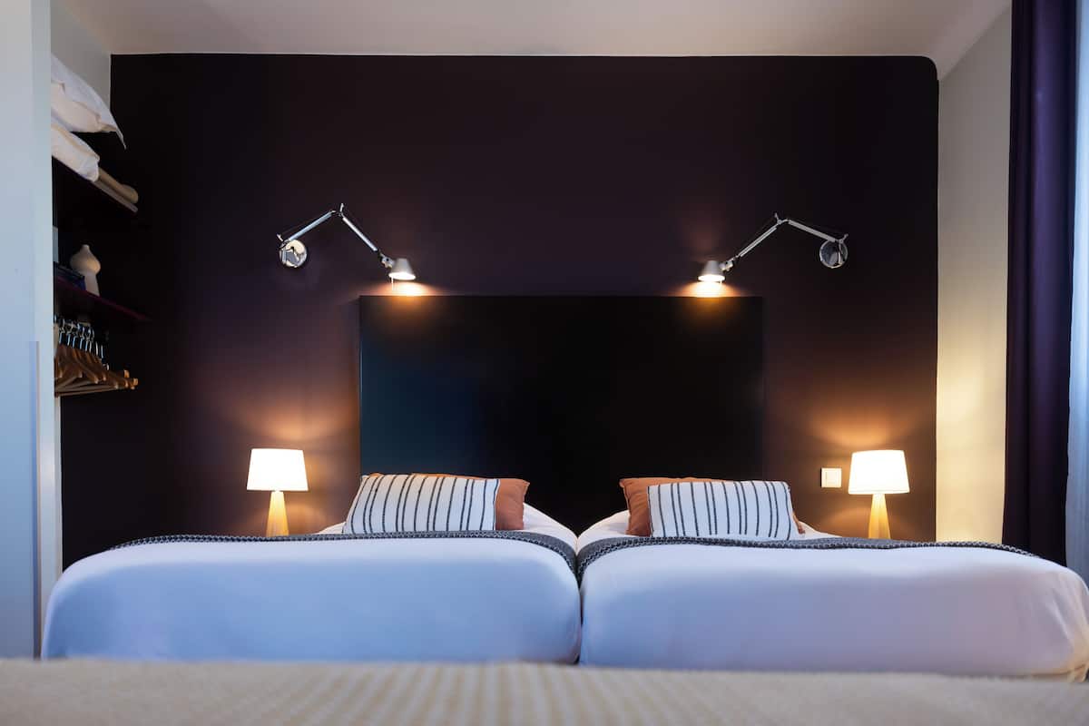 A double bed with two bedside lamps, a dark headboard, and a nightstand with a lamp.